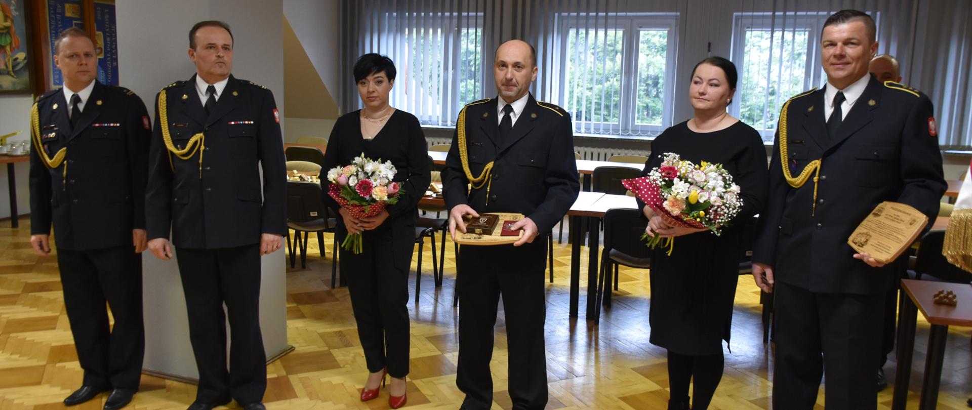 Zdjęcie przedstawia świetlicę komendy. Od lewej stoją Zastępca Dowódcy Jednostki Ratowniczo-Gaśniczej w Łańcucie młodszy brygadier Piotr Glac, następnie Zastępca Komendanta Powiatowego Państwowej Straży Pożarnej starszy brygadier Jacek Rejman, aspirant Dariusz Nicpoń wraz z żoną oraz aspirant sztabowy Janusz Witek wraz z żoną. Emeryci trzymają w rękach pamiątkowe ryngrafy zaś ich żony bukiety kwiatów.