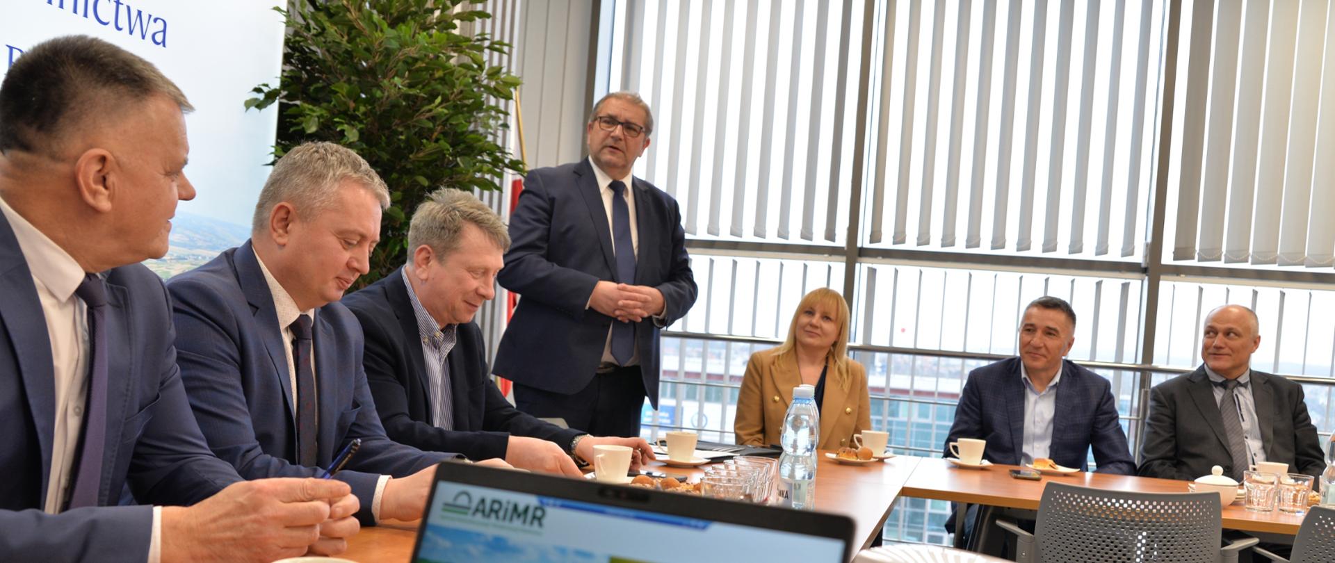 Grupa ludzi, sześciu mężczyzn i jedna kobieta. Jeden z mężczyzn stoi a reszta zajmuje miejsca siedzące przy stole. Wszyscy są ubrani elegancko w granatowe garnitury a kobieta w brązową marynarkę. Na pierwszym planie widać monitor laptopa na którym wyświetlona jest prezentacja. W pomieszczeniu są duże okna.