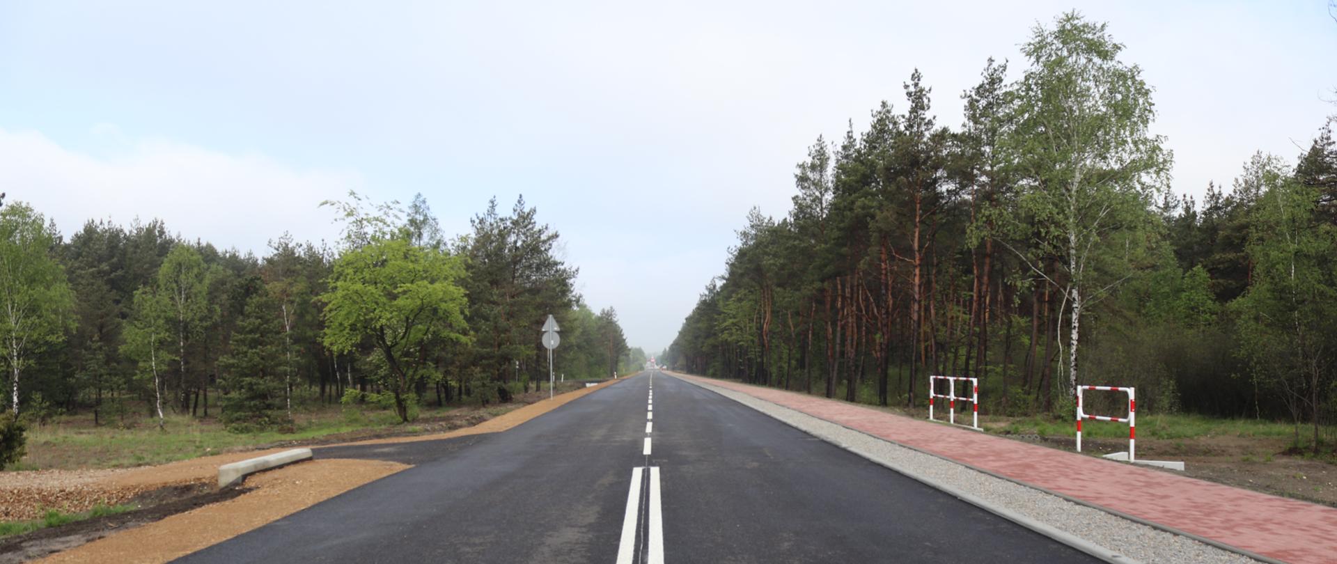 Na zdjęciu widać przebudowaną drogę, po prawej stronie jezdni znajduje się chodnik dla pieszych z czerwonej kostki. Po obu stronach jezdni rosną wysokie, zielone drzewa. 