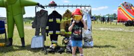 Chłopiec w hełmie strażackim, stoi na tle wystawy strażackich ubrań bojowych