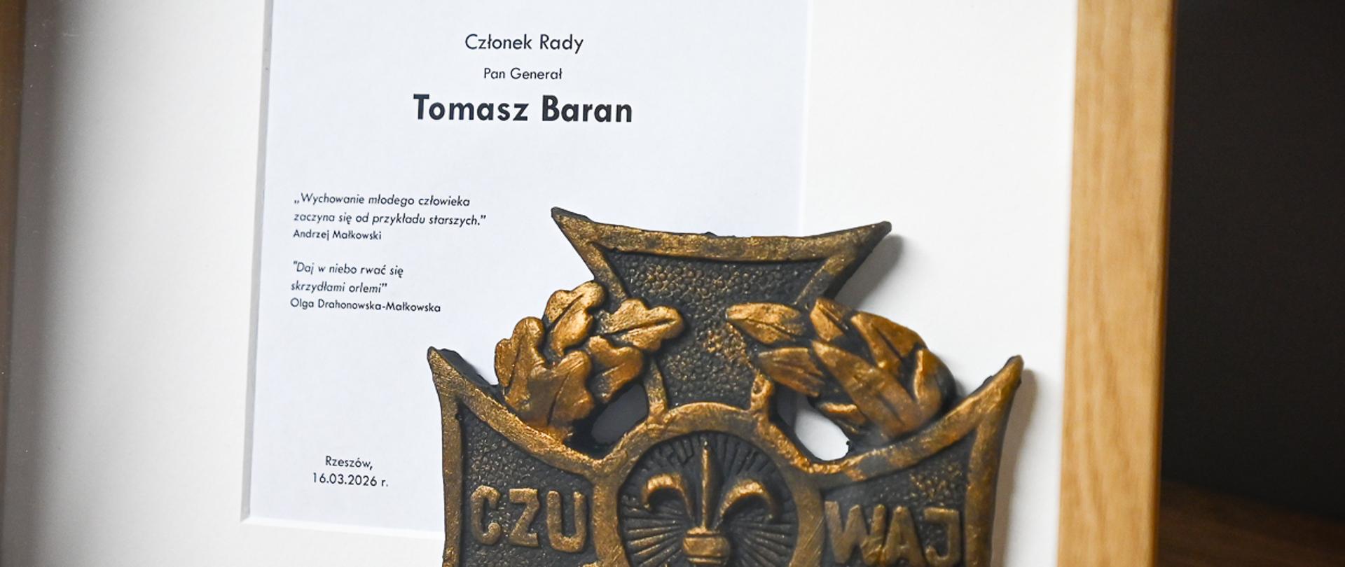 Zdjęcie przedstawia oprawiony w jasne drewno pamiątkowy zestaw. W górnej części znajduje się złota tabliczka z wygrawerowanym napisem: Podkarpacka Rada Przyjaciół Harcerstwa Chorągiew Podkarpacka ZHP im. Olgi i Andrzeja Małkowskich. Poniżej, na białym tle, umieszczono zadrukowaną kartę. Widnieje na niej tekst: Członek Rady Pan Generał Tomasz Baran „Wychowanie młodego człowieka zaczyna się od przykładu starszych.” Andrzej Małkowski „Daj w niebo rwać się skrzydłami orlemi” Olga Drahonowska-Małkowska Rzeszów, 16.03.2026 r.. W dolnej części zdjęcia, na pierwszym planie, spoczywa duży, wykonany z brązu, harcerski Krzyż Zasługi, ze słowem „CZUWAJ” rozdzielonym centralną lilijką.