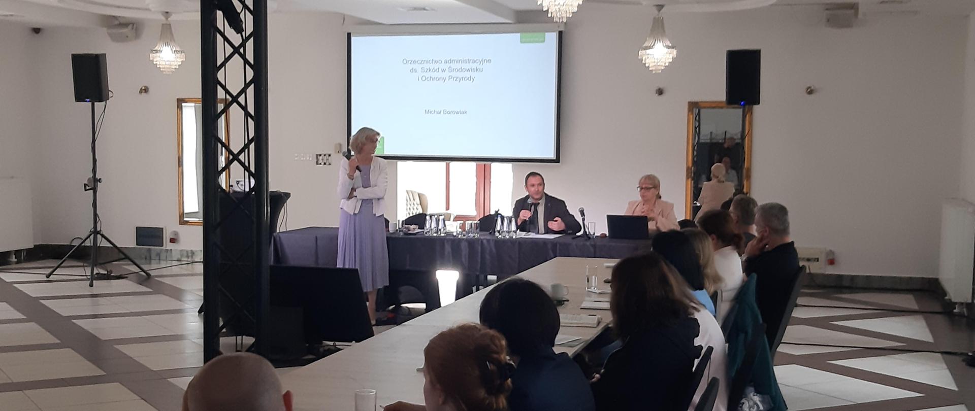 Widok dużej sali konferencyjnej. Przy stołach siedzą ludzie, którzy słuchają przemawiającego mężczyzny. Na ścianie wisi ekran na którym wyświetlana jest prezentacja pod tytułem: "Orzecznictwo administracyjne ds. Szkód w Środowisku i Ochrony Przyrody", autor Michał Borowiak.