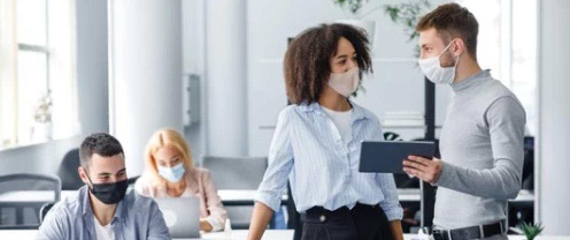 Na obrazku znajdują się cztery osoby. Dwie siedzą i pracują przy laptopach, a pozostali stoją i rozmawiają.