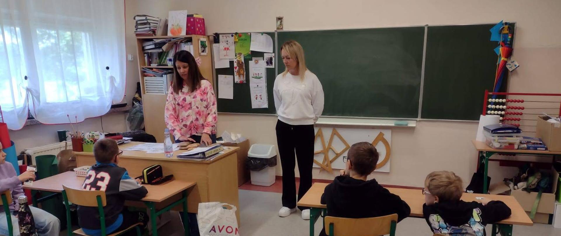 Dwie Panie Inspektor stojące w sali lekcyjnej, przed nimi w ławkach siedzą uczniowie