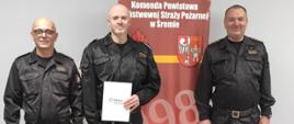 Na zdjęciu komendant powiatowy PSP w Śremie, wyróżniony funkcjonariusz oraz naczelnik pionu operacyjnego na tle baneru KP PSP Śrem