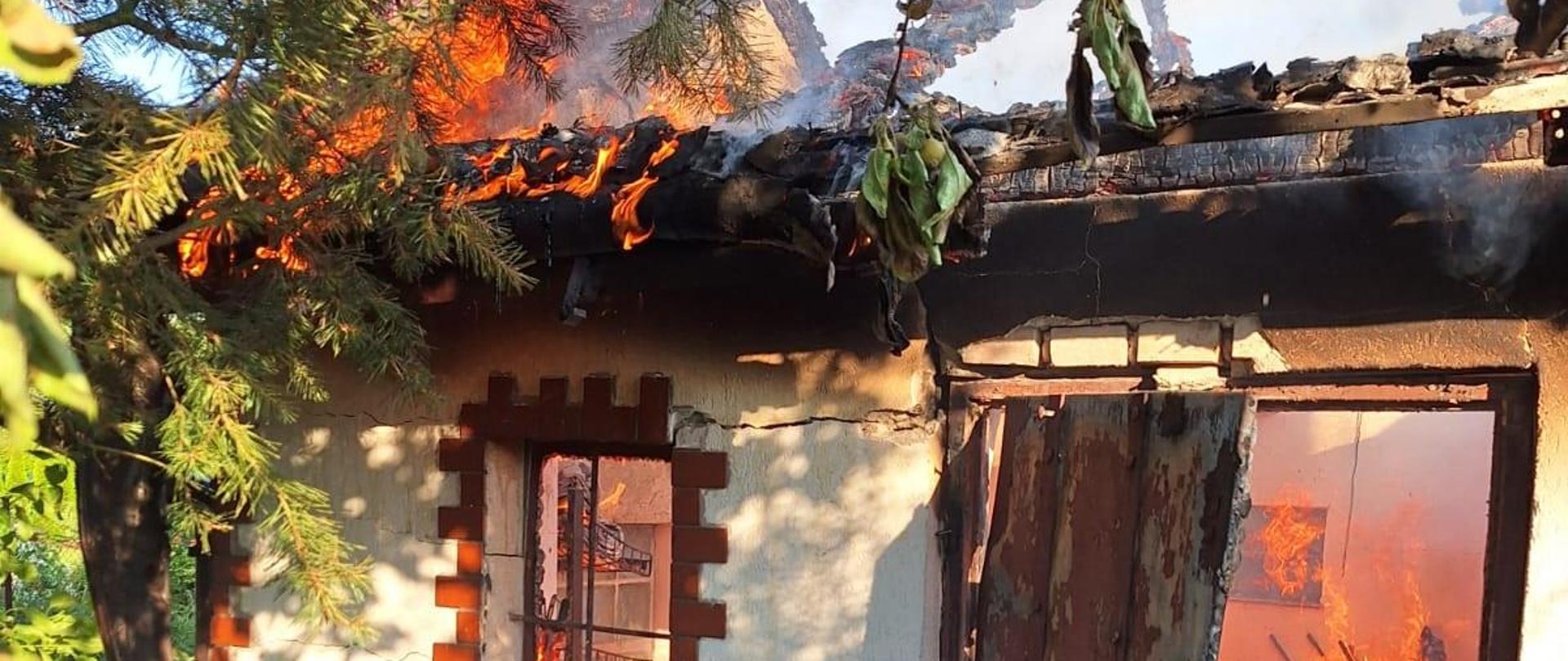 Biała altana ogrodowa, w oknach i drzwiach płomienie, spalony dach. Zielone drzewa i trawa.