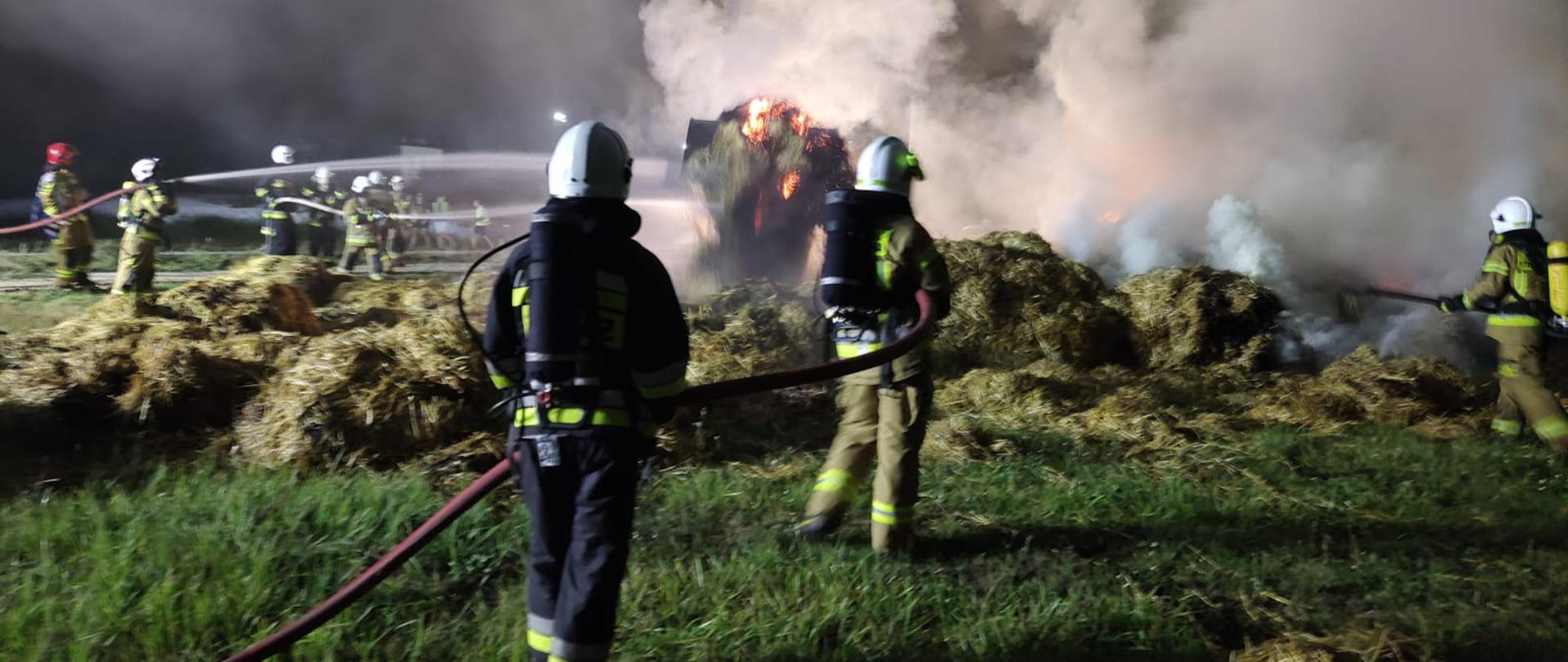 Na zdjęciu palace się baloty słomy i gaszący ten pożar strażacy na każdej lini gaśniczej po 2 strażaków widoczne 3 linie.
