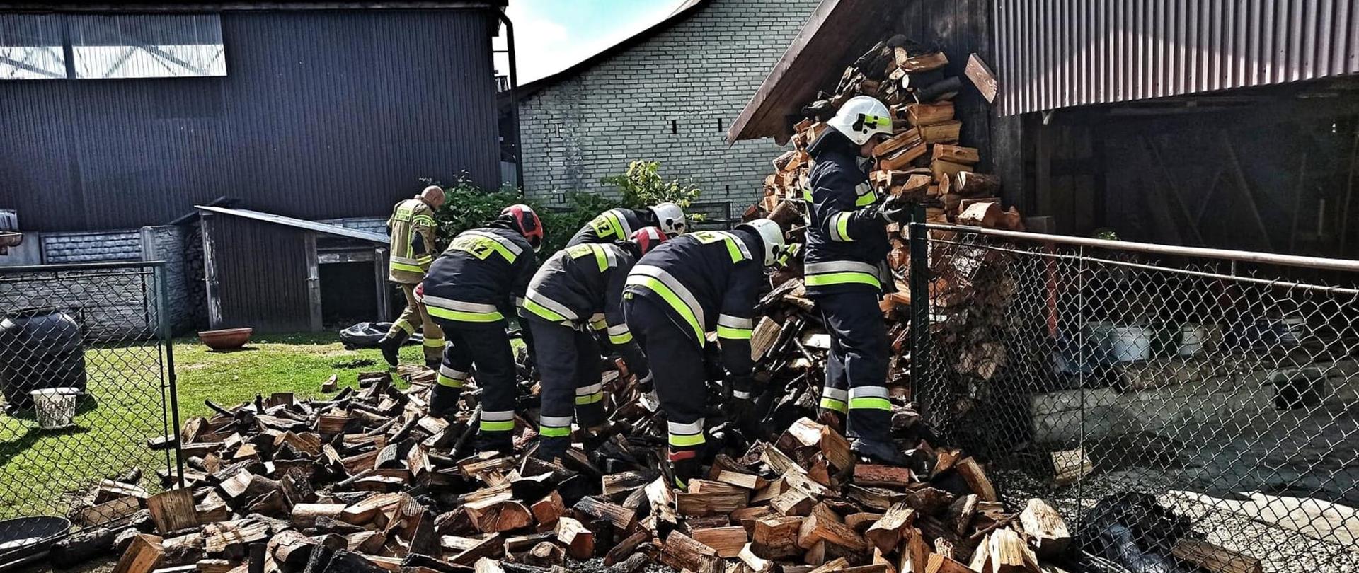 Na fotografii widocznych jest sześciu strażaków ubranych w mundury i hełmy. Stoją oni przy płocie i pochyleni są podczas przerzucania kawałków drewna opałowego z dużej pryzmy. Pryzma sprawia wrażenie jakby się przewróciła a jej część widoczna jest przy ścianie budynku umieszczonego za płotem. Prawdopodobnie drewno było składowane wzdłuż ściany budynku tak aby wyschło. Niektóre kawałki są nadpalone. W tle widoczne dwa duży obiekty budowlane - lewy ze ścianami z blachy falistej, prawy z białej cegły. Zdjęcie wygląda tak jakby było robione na podwórku.
