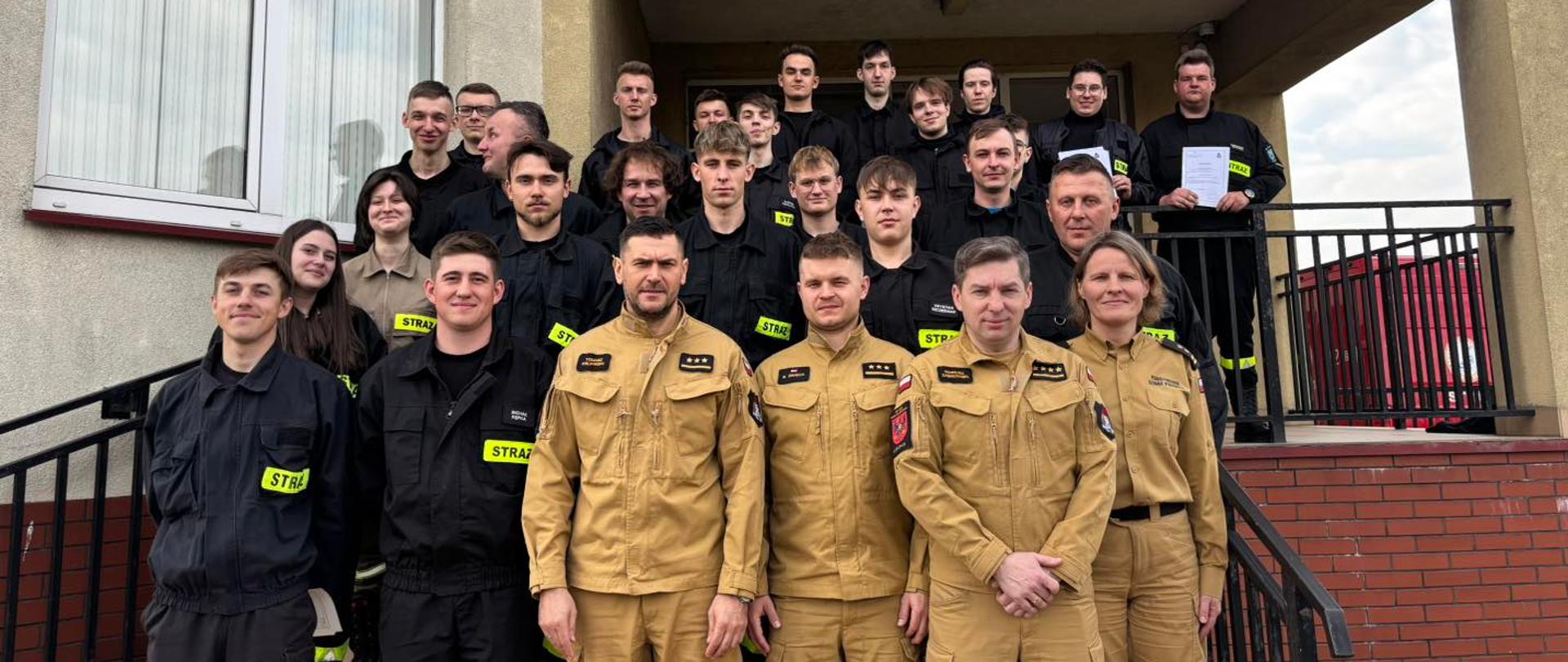 Na schodach zewnętrznych, prowadzących do drzwi wejściowych do komendy, kolejno na stopniach ku górze stoją druhowie OSP, którzy ukończyli szkolenie podstawowe, ubrani w umundurowanie koszarowe. Na samym dole stoi komisja egzaminacyjna, składająca się z oficerów Komendy Powiatowej Straży Pożarnej w Rawie Mazowieckiej.