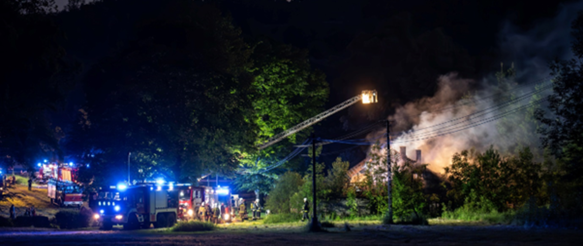 Z prawej strony zdjęcia widać spalony budynek, z którego unosi się para wodna. Z lewej strony zdjęcia znajdują się wozy strażackie. Nad budynkiem rozłożona jest drabina mechaniczna, z której oświetlane jest miejsce zdarzenia.