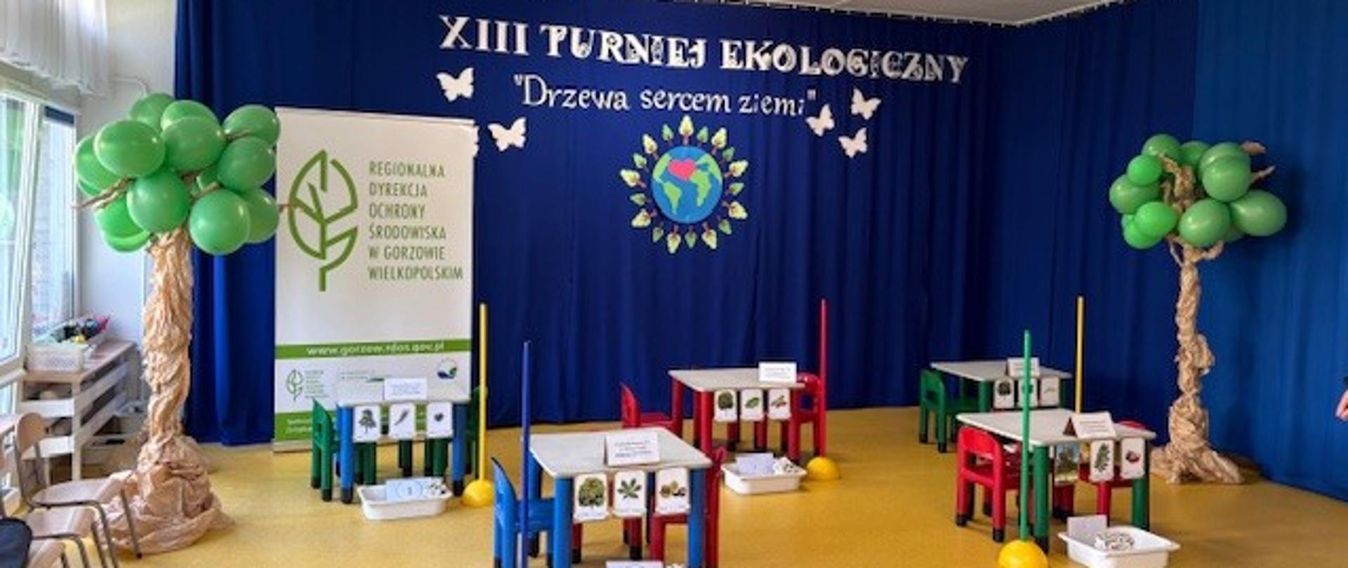 Na zdjęciu z lewej strony drzewo z papieru i zielonych balonów, pośrodku stoją kolorowe krzesełka i stoliki, po prawej stronie również stoi drzewo z papieru i zielonych balonów. W tle baner na niebieskim tle z napisem XIII Turniej Ekologiczny Drzewa Sercem Ziemi oraz baner Regionalnej Dyrekcji Ochrony Środowiska w Gorzowie Wielkopolskim