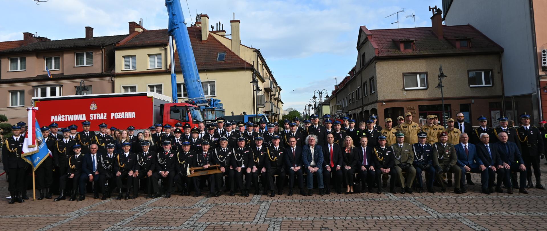 W centralnej części siedzą i stoją rzędy strażaków w czarnych mundurach galowych, funkcjonariusze w mundurach piaskowych oraz zaproszeni goście w strojach cywilnych i mundurach innych służb. Po lewej stronie widoczny jest poczet sztandarowy. W tle, za zgromadzonymi, stoi duży czerwony samochód kwatermistrzowski z napisem „Państwowa Straż Pożarna” oraz niebieski dźwig z uniesionym ramieniem. Całość kompozycji dopełniają zabytkowe kamienice.