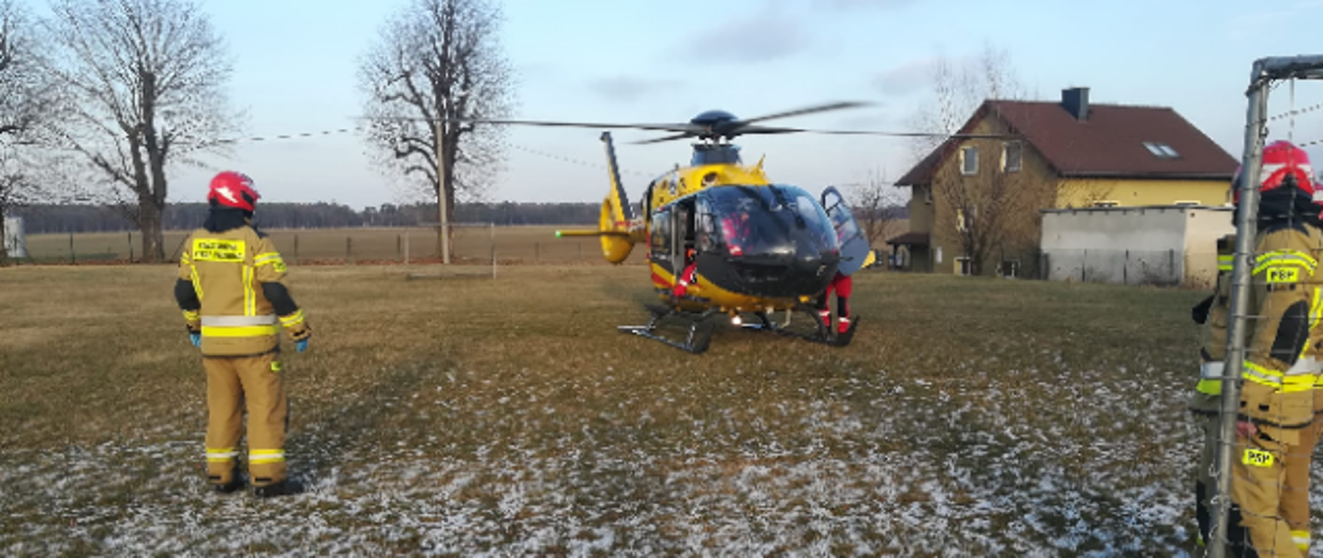 Zdjęcie przedstawia śmigłowiec LPR dwóch strażaków na tle domu