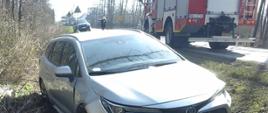Samochód osobowy marki Toyota po zderzeniu z samochodem ciężarowym stojący w przydrożnym rowie. W tle samochód ratowniczo-gaśniczy.