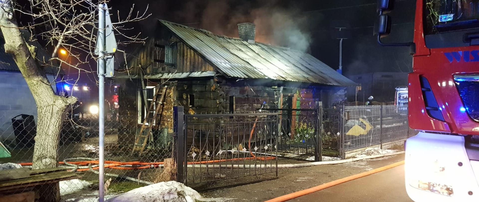 drewniany budynek z,którego wydobywa się dym. Obok widać częściowo samochód strażacki