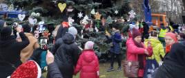 Osoby cywilne wraz z dziećmi chodzą wokół dużej ustrojonej choinki. W tle widoczne osoby na podnośniku sypiące śnieg.