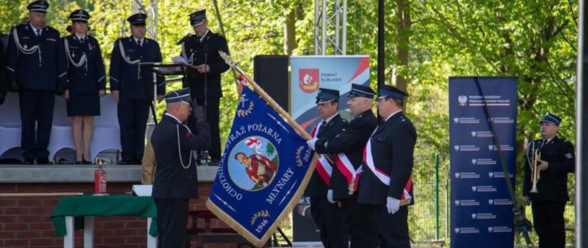 Podczas uroczystości strażacy w galowych mundurach stoją w szeregu przed sceną. Jeden z nich trzyma niebieski sztandar Ochotniczej Straży Pożarnej w Miłakowie, a obok stoją funkcjonariusze w białych rękawiczkach i biało‑czerwonych szarfach. Na scenie za nimi znajdują się kolejni mundurowi oraz mównica