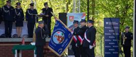 Podczas uroczystości strażacy w galowych mundurach stoją w szeregu przed sceną. Jeden z nich trzyma niebieski sztandar Ochotniczej Straży Pożarnej w Miłakowie, a obok stoją funkcjonariusze w białych rękawiczkach i biało‑czerwonych szarfach. Na scenie za nimi znajdują się kolejni mundurowi oraz mównica