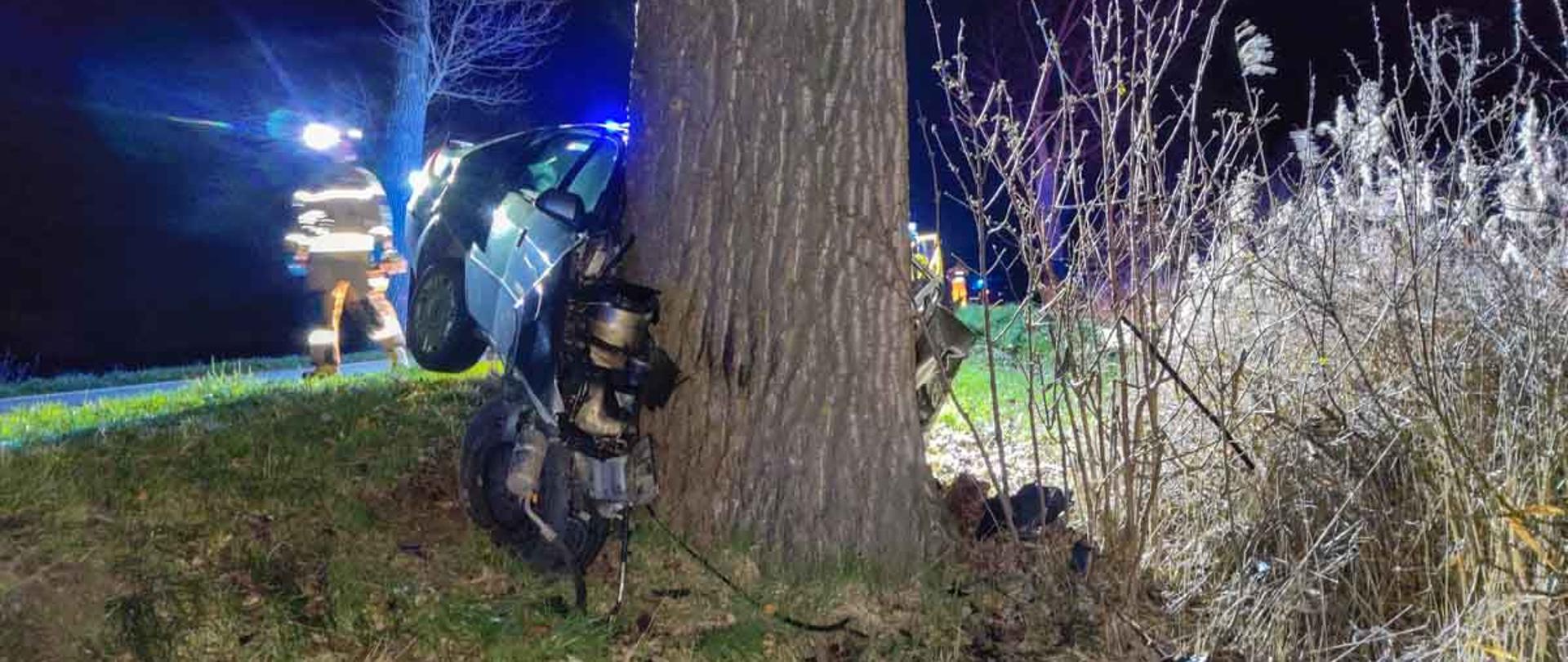 Na zdjęciu wrak pojazdu po uderzeniu czołowym w drzewo, obok przechodzący strażak w ubraniu koloru żółtego.