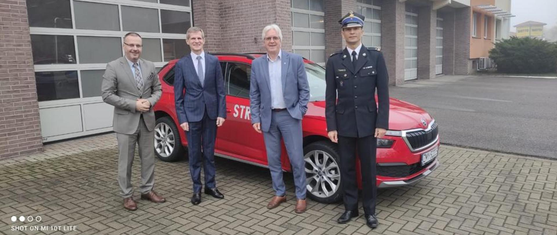 Na zdjęciu 4 mężczyzn obok oznakowanego samochodu osobowego w kolorze czerwonym