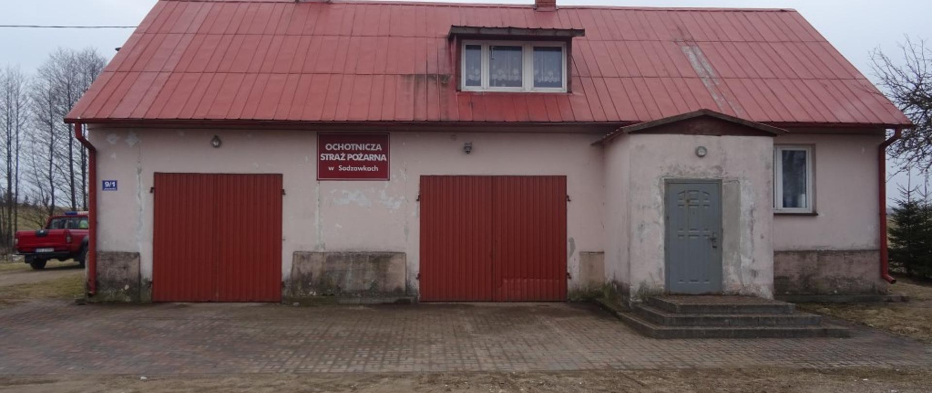 Budynek koloru szarego z dwoma bramami garażowymi koloru czerwonego. Wejście do budynku z prawej strony. Dach kryty blachą koloru czerwonego. Podjazd wyłożony kostką brukową. 