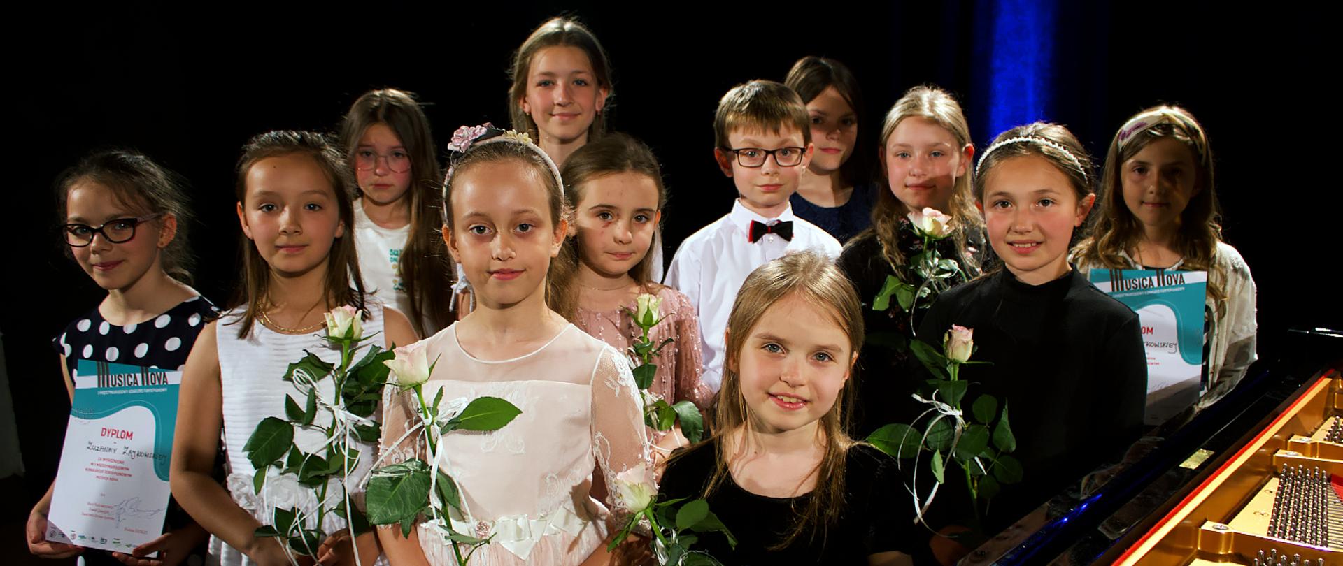 Na zdjęciu grupa 12 dzieci, 11 dziewczynek i jeden chłopiec, dzieci stoją, patrzą w kierunku robiącego zdjęcie, w rękach trzymają białe róże i dyplomy, z prawej strony widać fragment fortepianu.