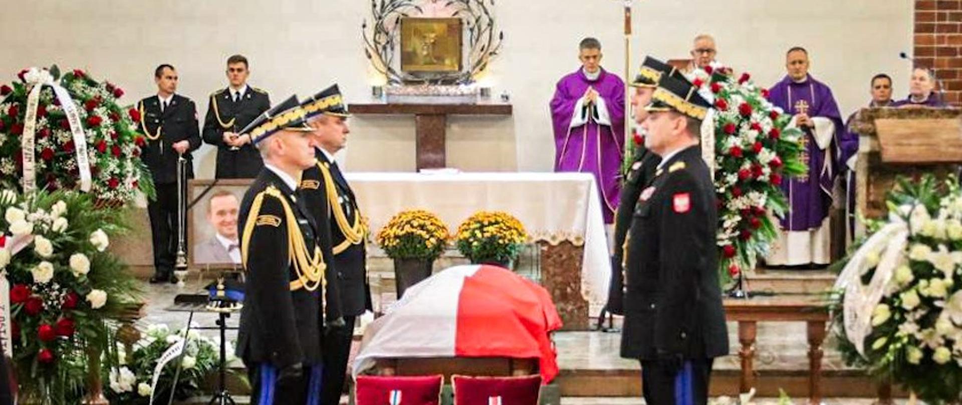 Przy trumnie odzianej w Polską flagę wartę pełnią generałowie straży pożarnej. Przed trumną na poduszkach są medale. Dookoła wieńce pogrzebowe. 