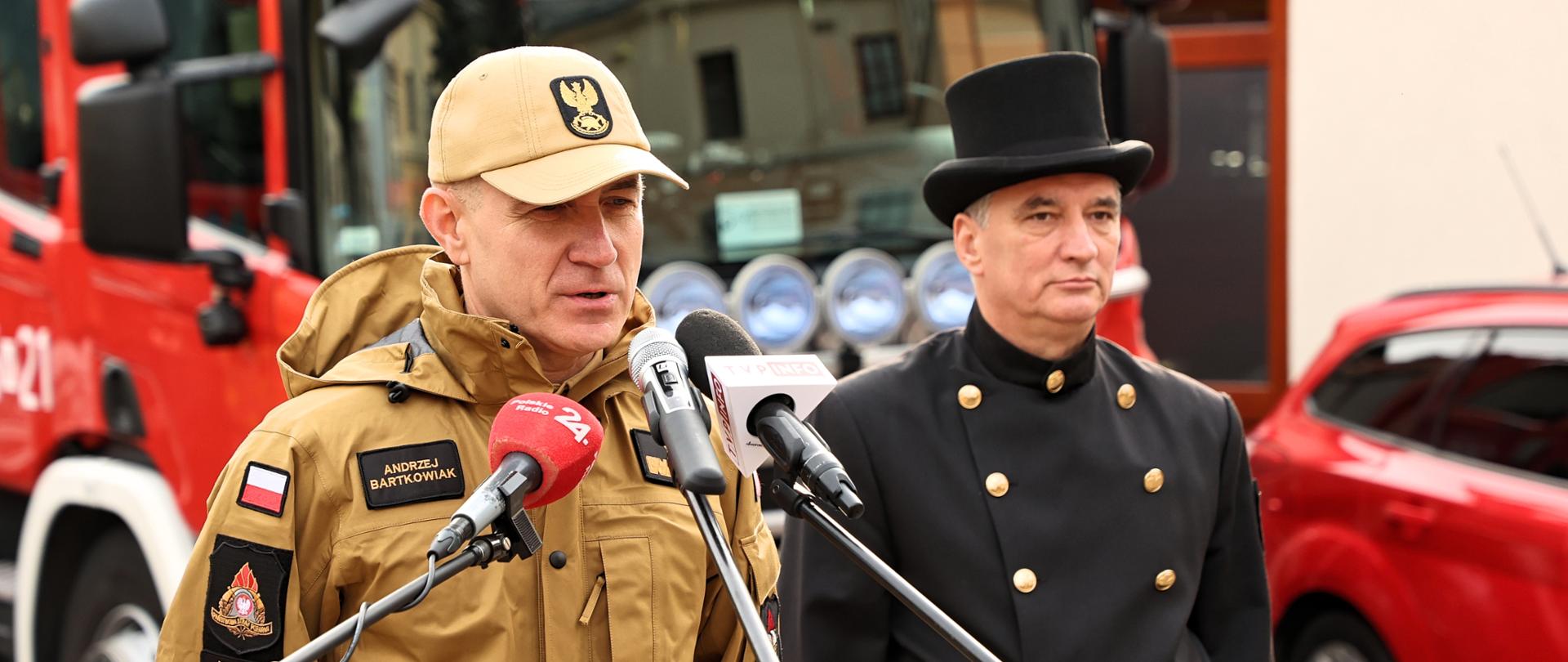 Na zdjęciu komendant główny w umundurowaniu służbowym i kominiarz w swoim stroju służbowym. stoją przy mikrofonie podczas briefingu. W tle pojazdy pożarnicze.