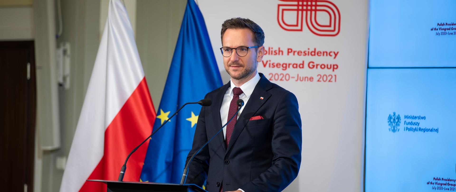 Na zdjęciu wiceminister funduszy i polityki regionalnej Waldemar Buda przemawiający w trakcie XIV Międzynarodowej Konferencji Ewaluacyjnej. W tle flaga Polski i Unii Europejskiej oraz baner "Polska Prezydencja w Grupie Wyszehradzkiej lipiec 2020 - czerwiec 2021".