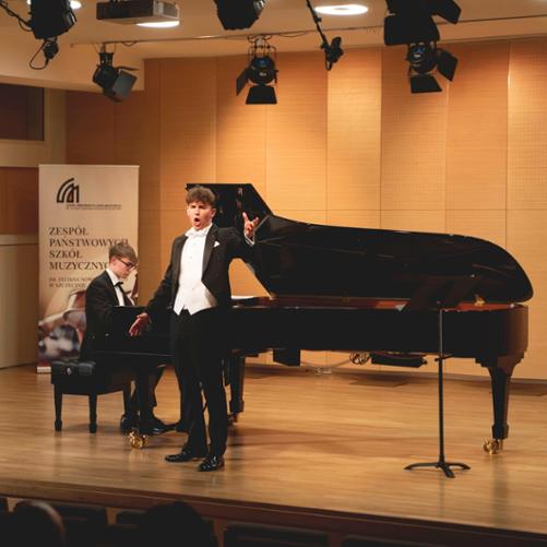 zdjęcie przedstawia ucznia śpiewającego z towarzyszeniem pianisty grającego na otwartym fortepianie na scenie sali koncertowej, w tle baner szkoły, inne ujęcie z szerszej perspektywy