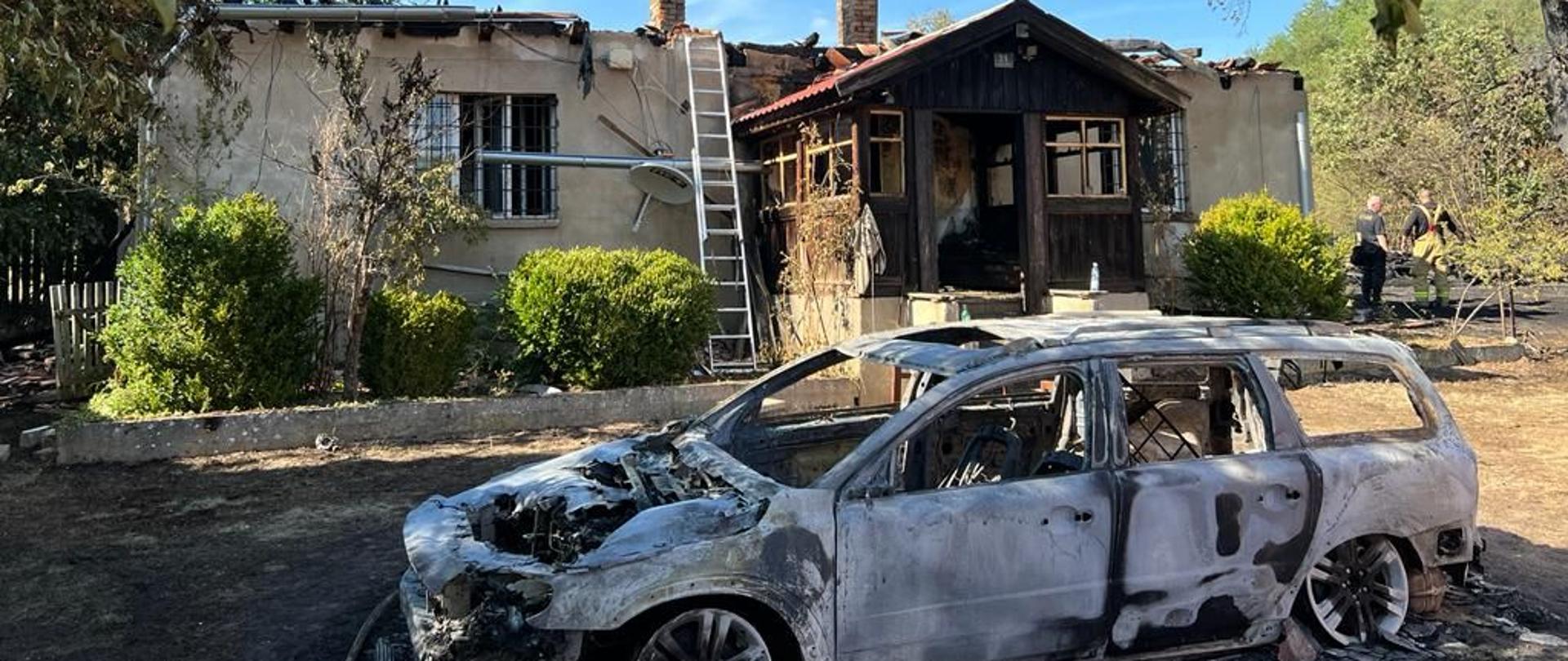 Na pierwszym planie spalony samochód osobowy, w tle budynek mieszkalny po pożarze.