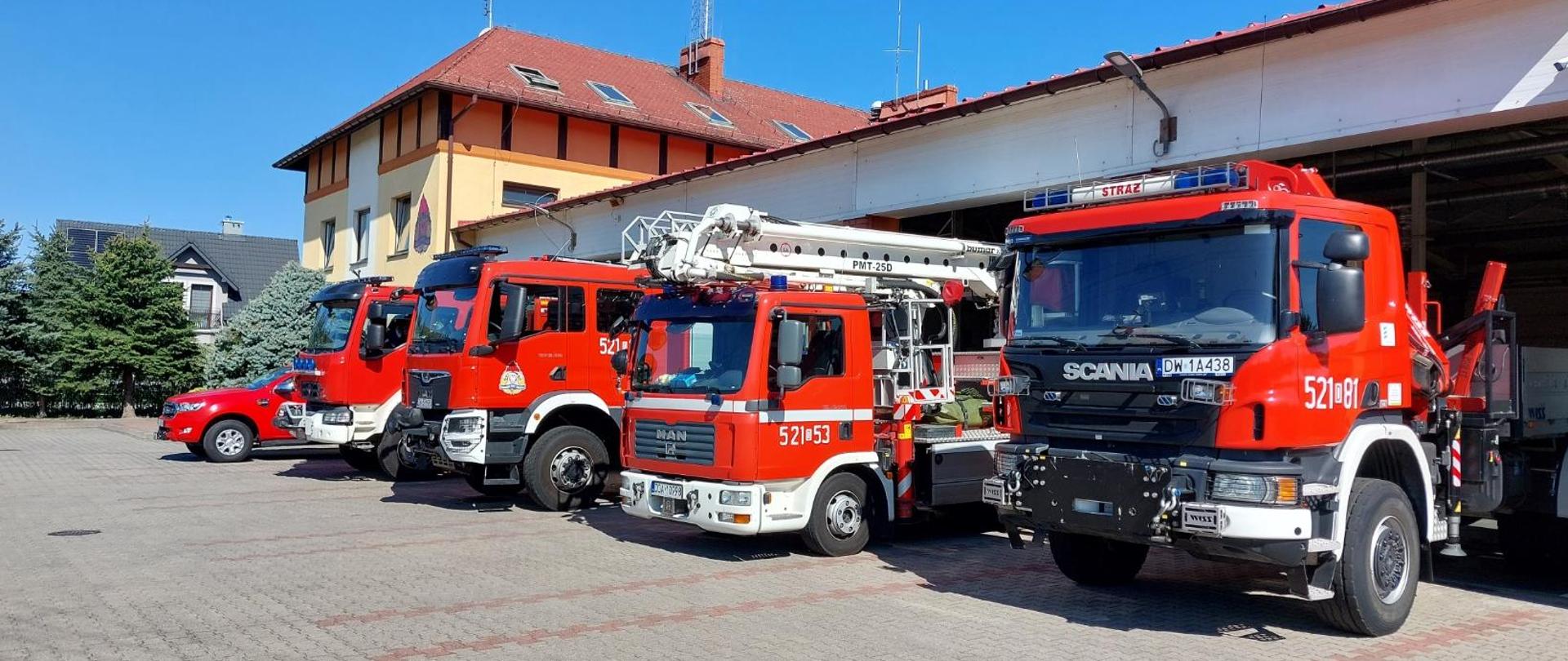 Zdjęcie przedstawia plac manewrowy przed garażami jednostki ratowniczo-gaśniczej, na którym stoi w rzędzie pięć czerwonych pojazdów pożarniczych oraz jeden mniejszy pojazd pomocniczy.
Od prawej strony widoczne są: ciężki samochód kwatermistrzowski Scania, samochód specjalny z podnośnikiem hydraulicznym MAN, dwa średnie lub ciężkie samochody ratowniczo-gaśnicze MAN oraz Renault oraz czerwony samochód rozpoznawczo-ratowniczy typu pick-up. W tle znajduje się murowany budynek komendy z masztami antenowymi na dachu. Zdjęcie wykonano w słoneczny dzień przy bezchmurnym niebie.
