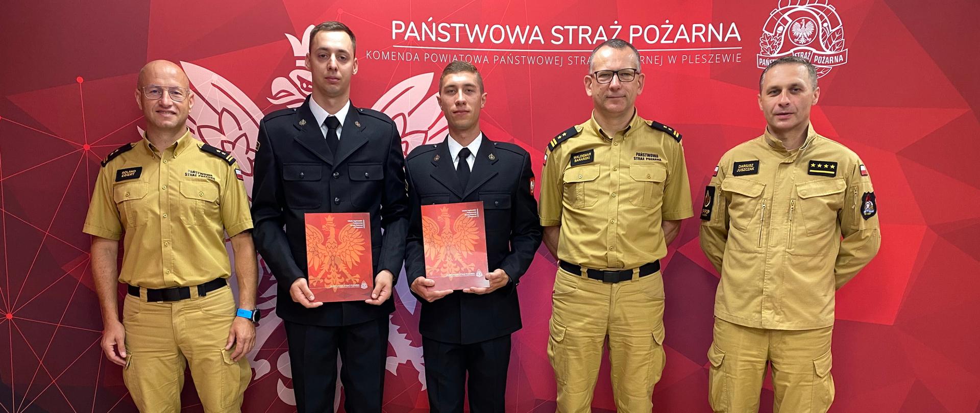 Pięciu mężczyzn ubranych w mundury Państwowej Straży Pożarnej stoi w szergu, dwóch z nich - w mundurach wyjściowych trzyma przed sobą dyplomy, w tle widoczny czerwony baner z logiem i napisem Państwowa Straż Pożarna