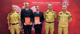Pięciu mężczyzn ubranych w mundury Państwowej Straży Pożarnej stoi w szergu, dwóch z nich - w mundurach wyjściowych trzyma przed sobą dyplomy, w tle widoczny czerwony baner z logiem i napisem Państwowa Straż Pożarna