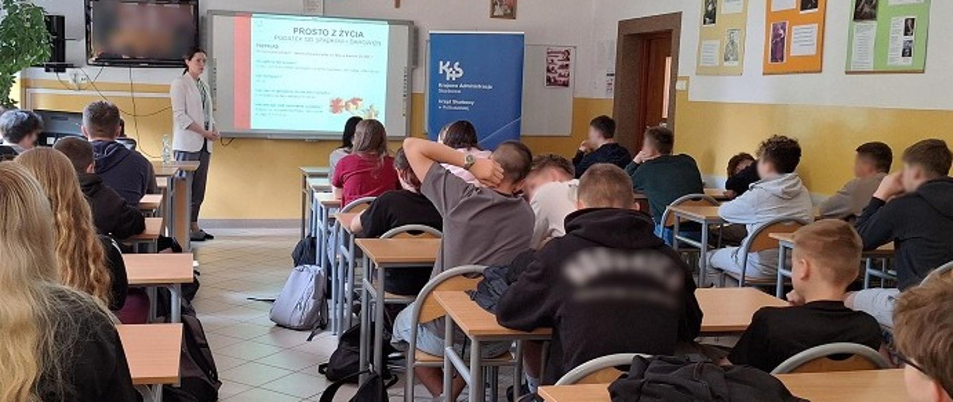 Uczniowie siedzą w ławkach szkolnych. Przed nimi stoi ekspertka. Na ekranie wyświetlana jest prezentacja.