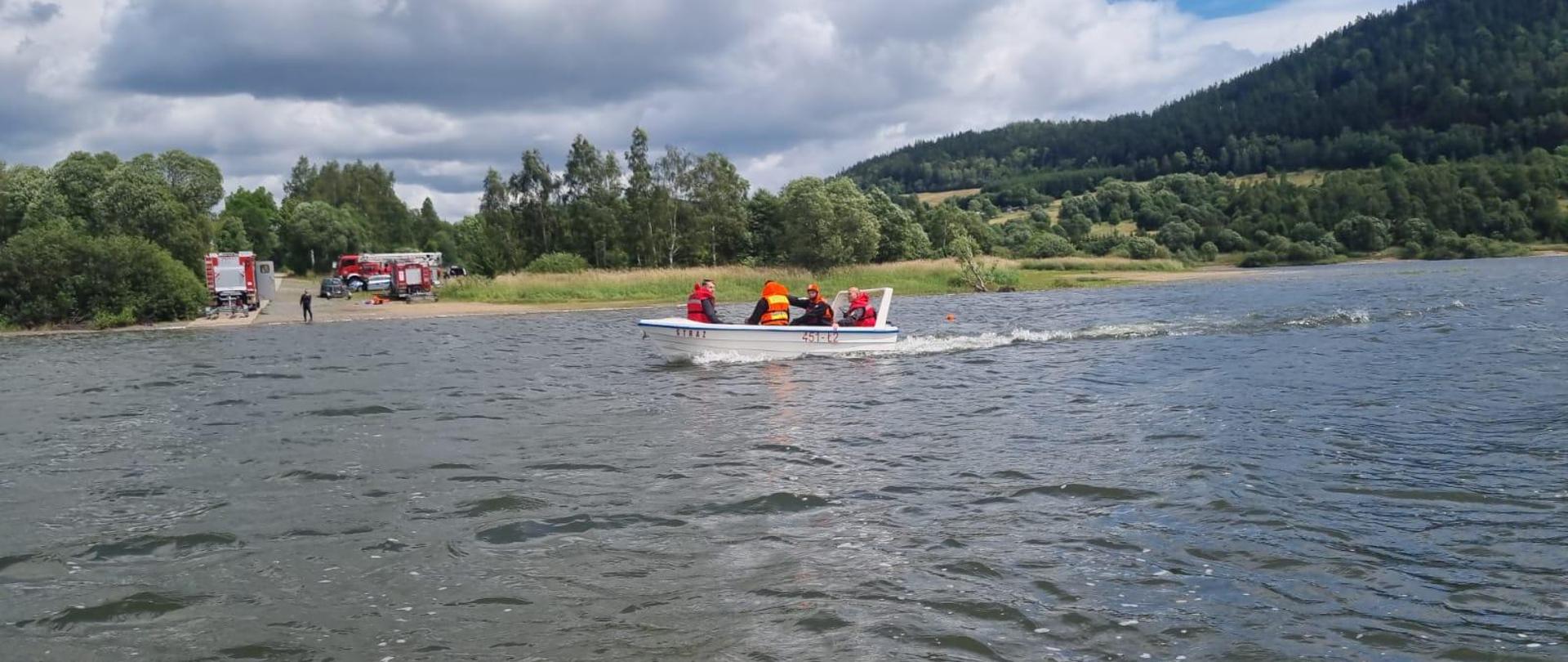 Plan główny łódź z ratownikami w tle dostrzegamy ustawione samochody ratowniczo gaśnicze na brzegu.