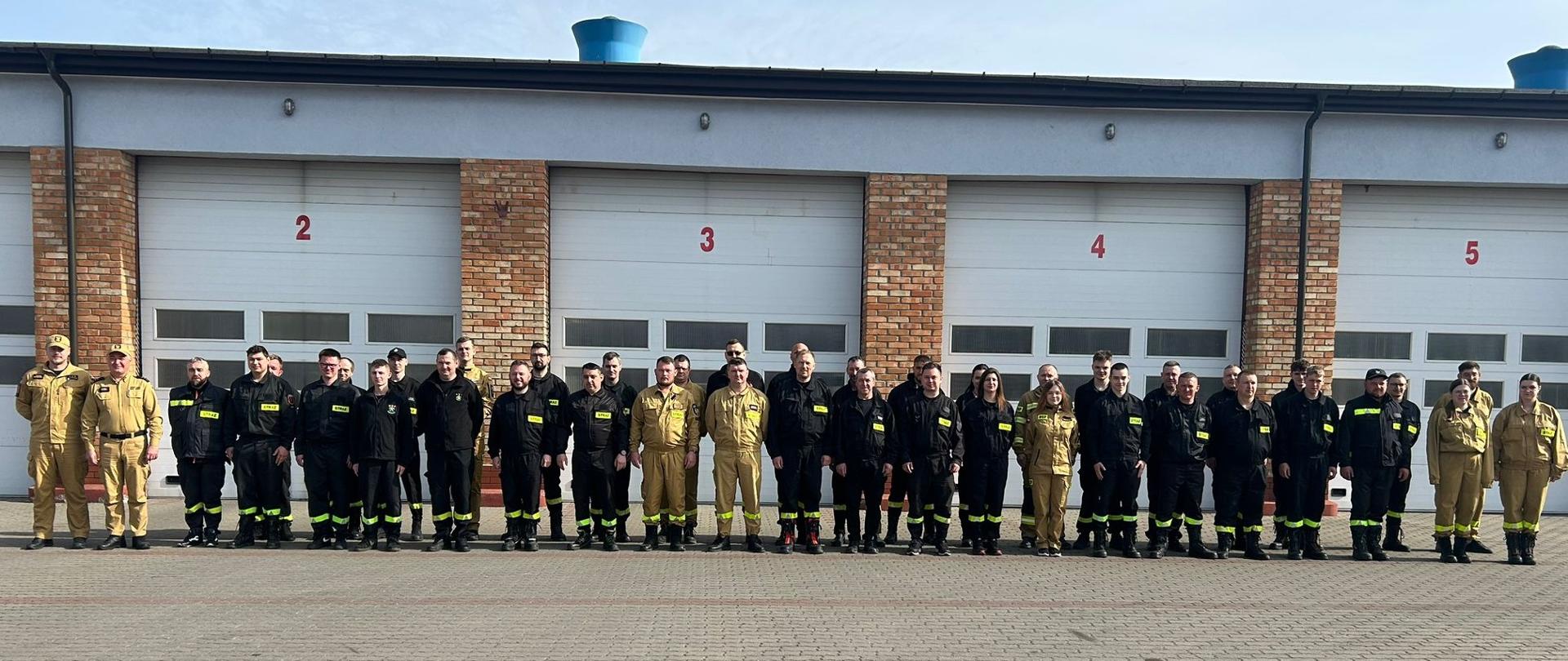 Pamiątkowe zdjęcie grupowe uczestników egzaminu szkolenia podstawowego strażaków ratowników OSP, wykonane przed garażami Komendy Miejskiej PSP w Ostrołęce. Kilkudziesięciu mężczyzn i kobiet stoi w jednym szeregu na placu manewrowym, ubranych w czarne oraz piaskowe mundury koszarowe. W tle widoczne są białe bramy garażowe z czerwonymi numerami od 2 do 5, oddzielone ceglanymi filarami.
