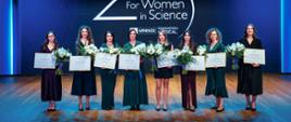 Na tle granatowej ściany z napisem 25 for Women in Science osiem elegancko ubranych kobiet, trzymają bukiety białych kwiatów i dyplomy w ramkach.