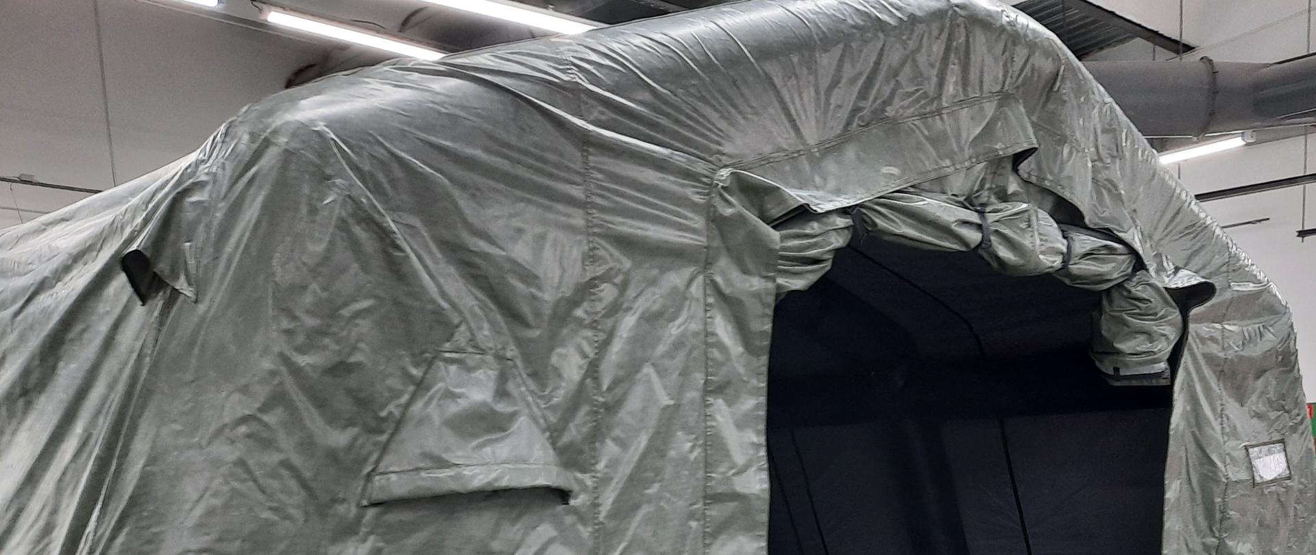 Pneumatyczny namiot ratowniczy w kolorze oliwkowej zieleni. Na zdjęciu widok przednio-boczny. Otwarte wejście, w którym widać wnętrze namiotu w kolorze grafitowym.