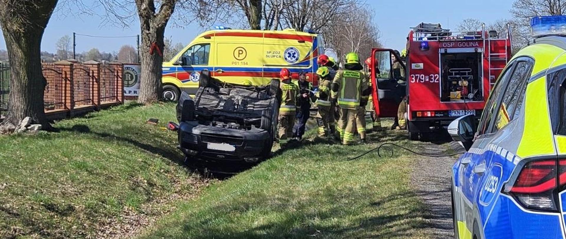 Samochód osobowy leży w przydrożnym rowie na dachu. Przy samochodzie stoją strażacy. Z tyłu widać samochód strażacki i karetkę pogotowia. Z przodu stoi radiowóz. 