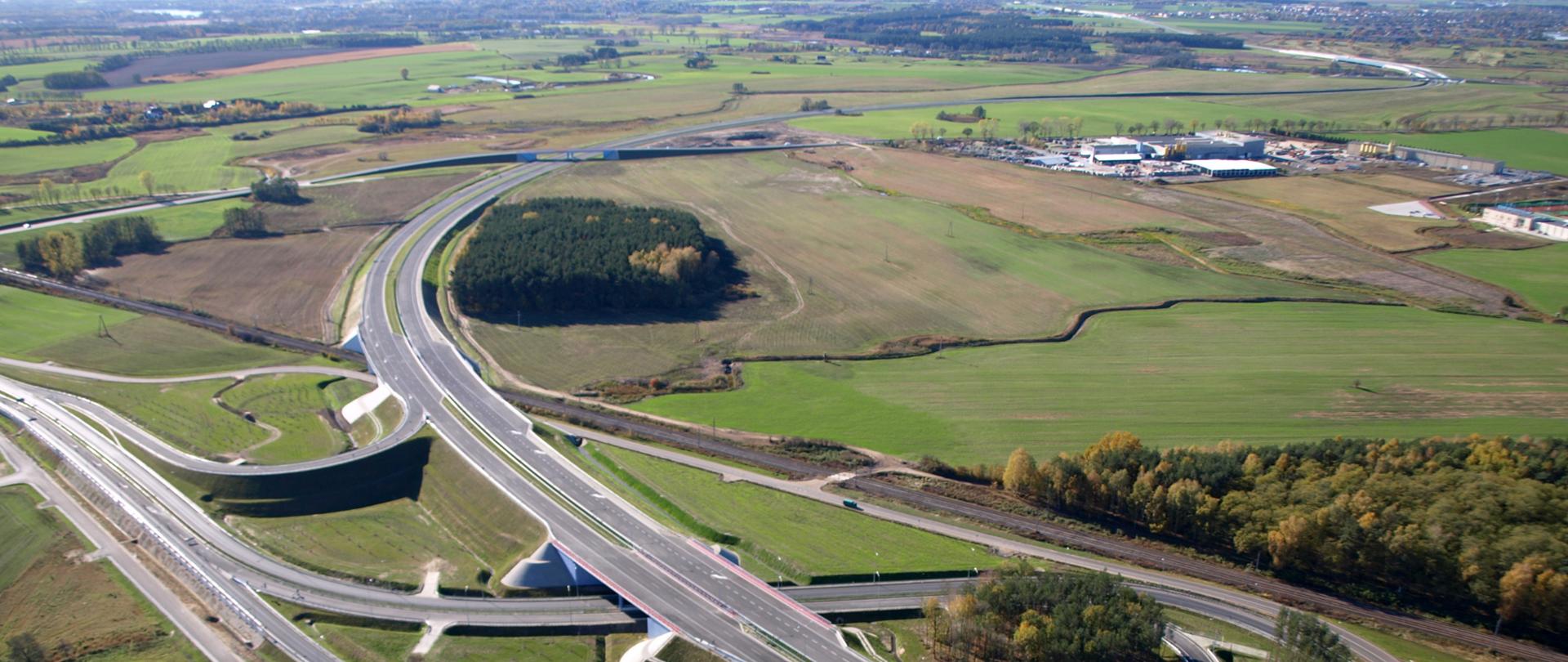 
Zdjęcie przedstawiające węzeł drogowy Poznań Północ na drodze ekspresowej S11. Węzeł widoczny z powietrza przebiegający między łąkami, polami i zabudową magazynową. Na jednej z łącznic doprowadzających ruch do węzła widoczne poruszające się pojazdy.