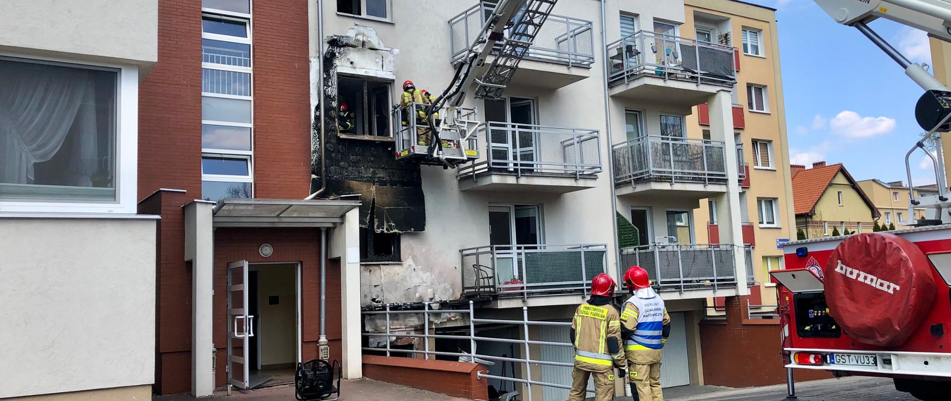 Zdjęcie przedstawia miejsce akcji, widać rozstawiony wysięgnik, wypalona elewacja budynku, na środku kierujący działaniami strażak