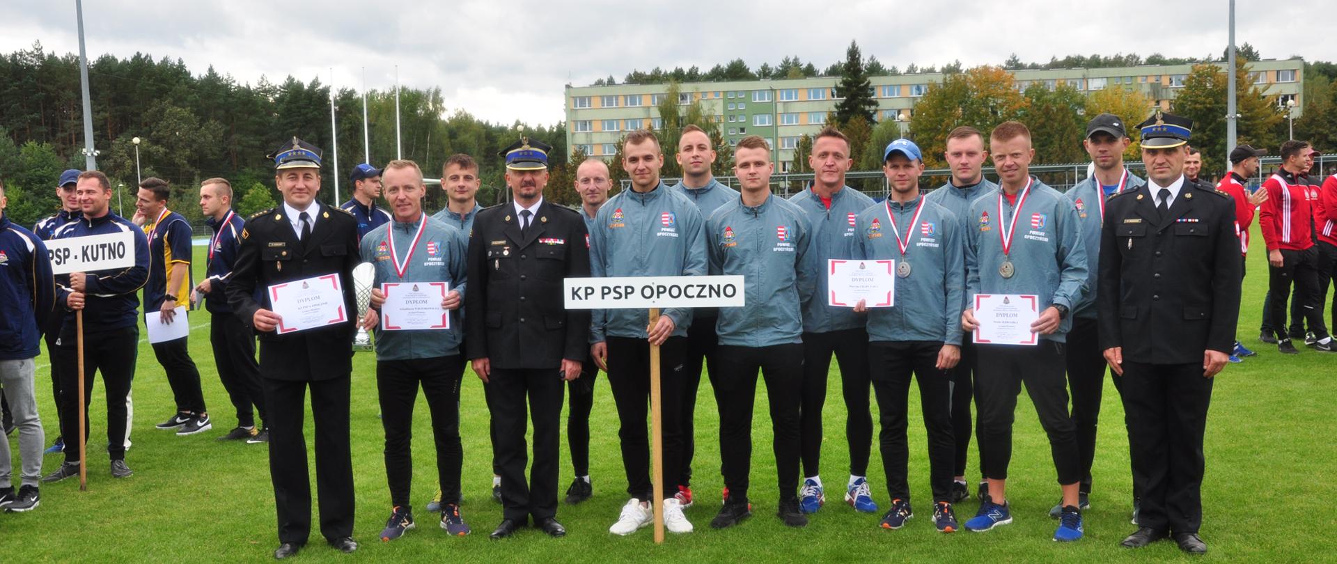 Drużyna strażaków z Komendy Powiatowej PSP Opoczno w szarych dresach trzymają dyplomy i tabliczkę ze skrótem komendy którą reprezentują. W śród nich komendanci w umundurowaniu wyjściowym. W tle zawodnicy z innych komend i budynek wielorodzinny.