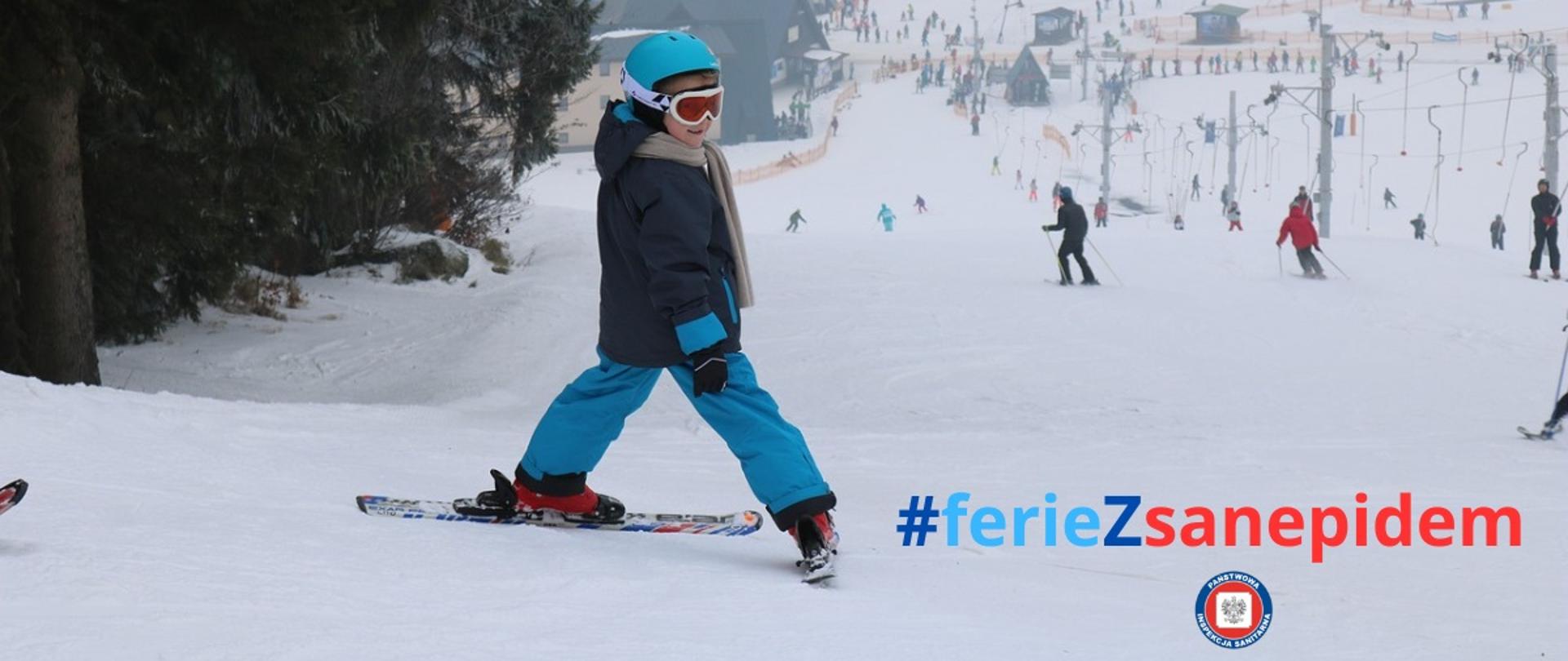 na szczycie stoku narciarskiego dziecko na nartach zwrócone w stronę odbiorcy zdjęcia, obok napis #feriezsanepidem