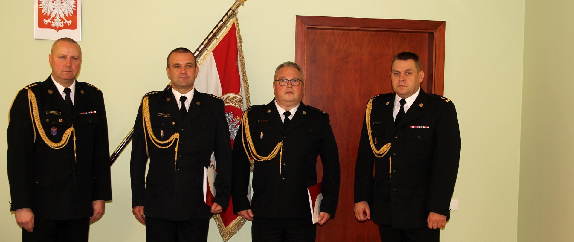 na tle zielonej ściany sztandaru oraz godła na ścianie stoi czterech strażaków na baczność ubranych w ciemne mundury ze złotym sznurem, 