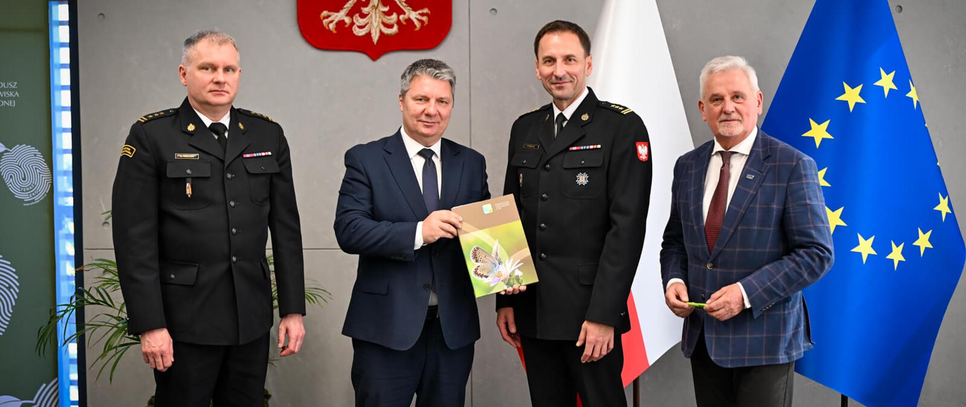 Czworo mężczyzn stoi w rzędzie w oficjalnej sali. W centrum mężczyzna w garniturze trzyma i pokazuje otwarty kolorowy folder z grafiką przyrodniczą mężczyźnie w mundurze strażackim. Za nimi widoczne są flagi Polski i UE oraz godło.
