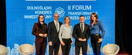 Zdjęcie przedstawia pięć osób, które stoją na scenie. W ich gronie znajduje się Sekretarz Stanu w Ministerstwie Rozwoju i Technologii, Michał Jaros oraz Łukasz Marciniak, Zastępca Dyrektora Departamentu Planowania Przestrzennego. W tle znajduje się ekran ledowym, na którym znajduje się napis: Dolnośląski Kongres Inwestycyjny oraz II Forum Transformacji Przemysłowej.