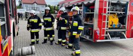 czterech strażaków ubranych w ubrania specjale stoi pomiędzy samochodami pożarniczym i przygotowując się do zaliczenia egzaminu praktycznego