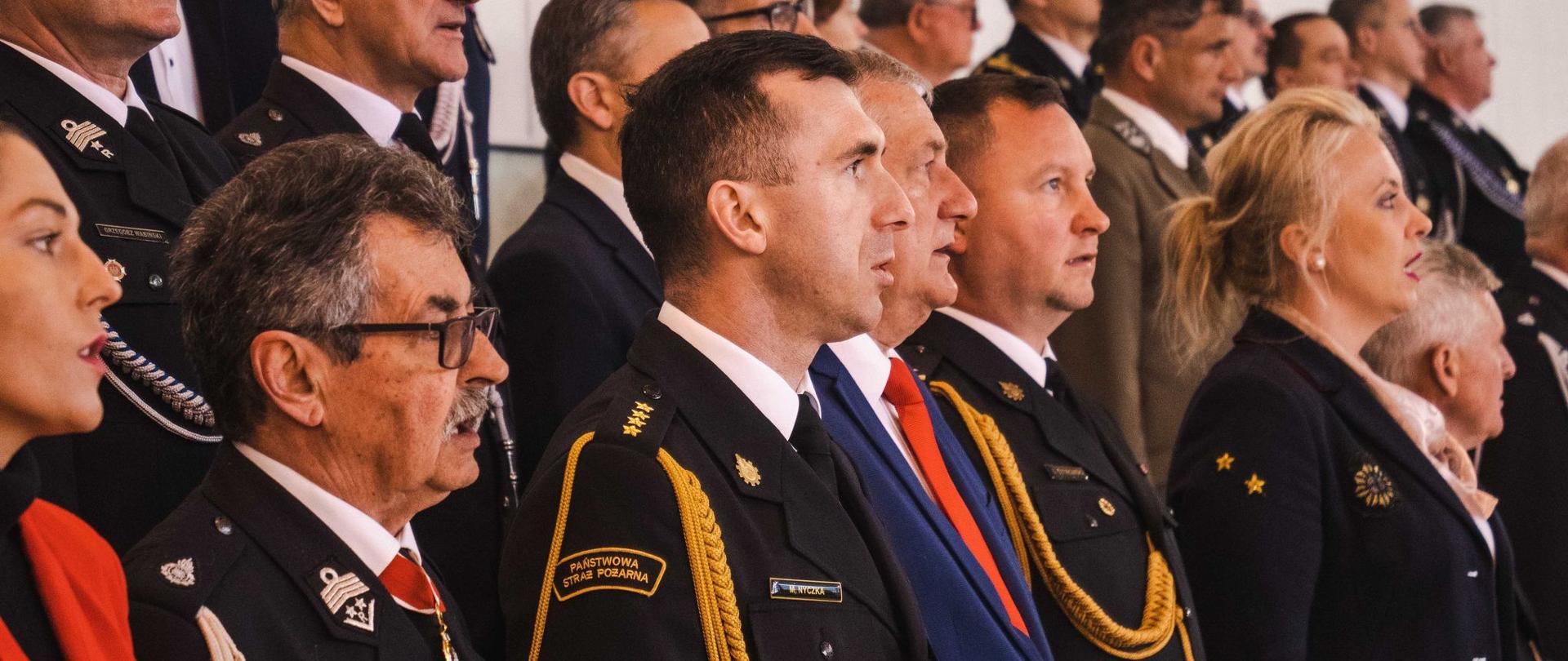 Zdjęcie przedstawia komendanta powiatowego oraz zaproszonych gości podczas uroczystości. Śpiewają oni hymn państwowy