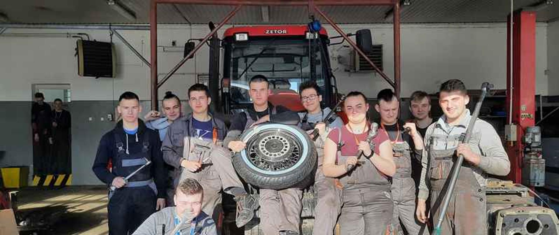 Na tle ciągnika rolniczego stoi grupa młodzieży ubranej w stroje robocze.